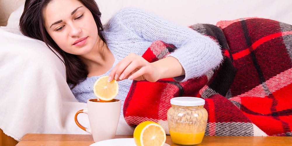Junge Frau liegt mit Decke auf der Couch vor ihr eine Tasse heiße Zitrone und Honig