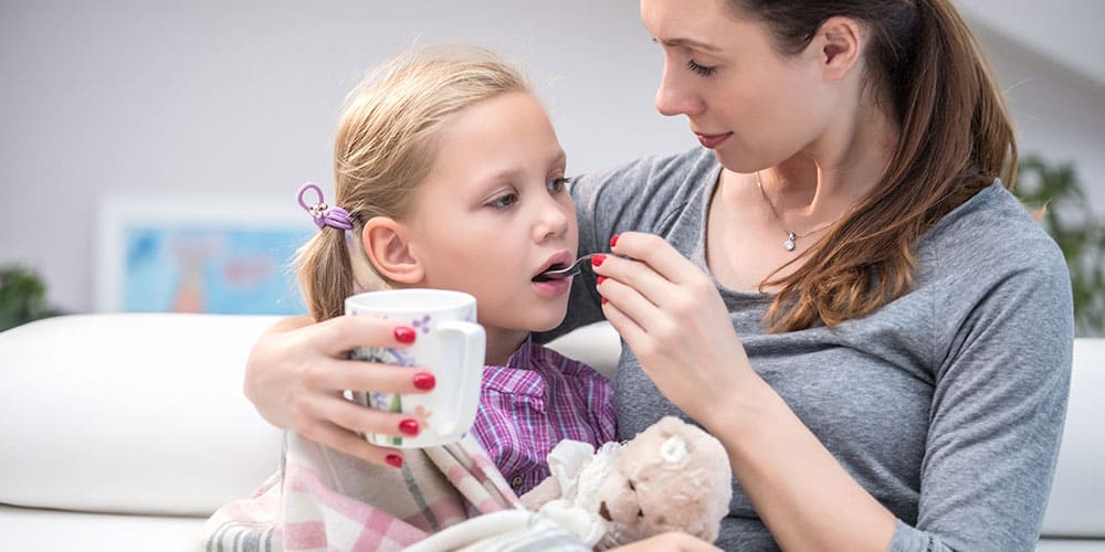 Mutter gibt ihrer Tochter einen Löffel mit Honig, in der Hand hält sie eine Tasse Tee