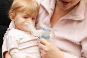 Kleinkind inhaliert auf dem Schoß seiner Mutter mit einem Inhalationsgerät