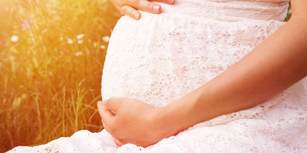 Bauch einer schwangeren Frau in weißem Kleid im Hintergrund eine Blumenwiese