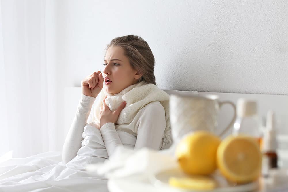 Junge Frau sitzt aufrecht im Bett und hustet- im Vordergrund ein paar Zitronen und eine Tasse Tee