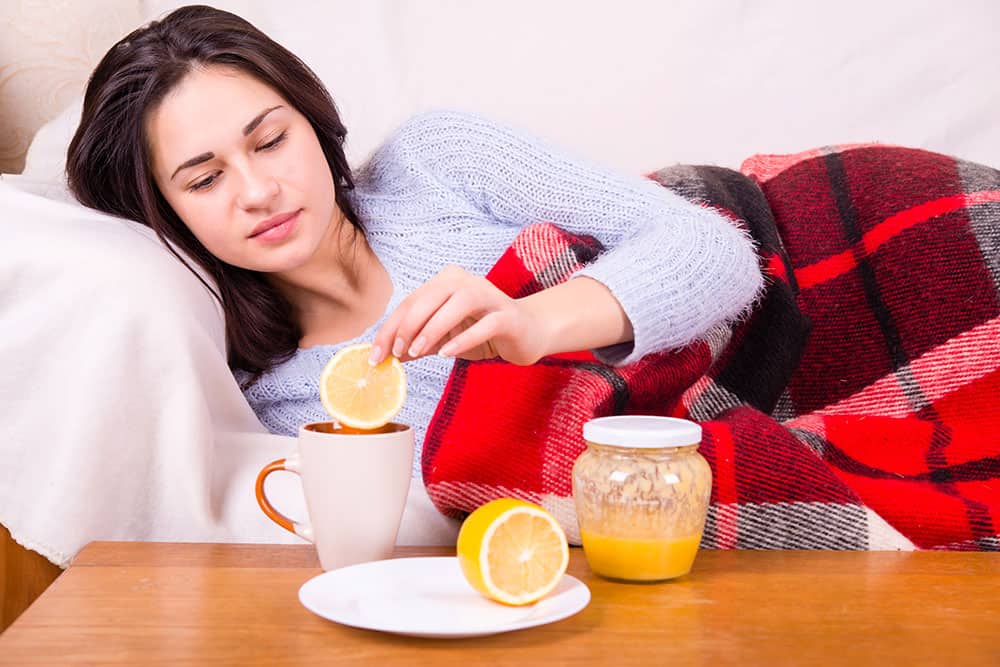 Junge Frau liegt mit einer Decke auf dem Sofa und macht eine schreibe Zitrone in eine Teetasse